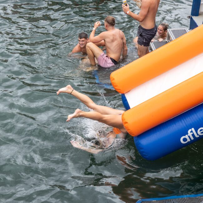 Several people are enjoying themselves playing in water next to a floating platform. One person is captured mid-air sliding off a vibrant inflatable water slide, while others are swimming near the slide or sitting on the platform, soaking up the sun.