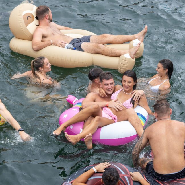 A group of people enjoying a swim in a body of water, with some floating on various inflatables like a donut, unicorn, and bear. People are smiling and laughing, indicating a cheerful and fun atmosphere under the sunny sky.