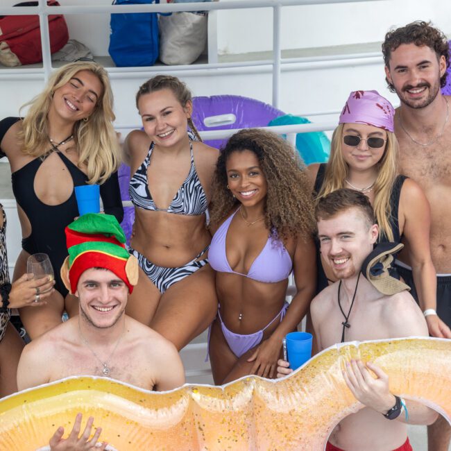 A group of eight people in swimwear smiling and posing for a photo on a boat. Some hold inflatable pool toys and plastic cups. There are festive accessories, like a Santa hat and elf hat, among the group, adding to the fun atmosphere. The background shows the boat's railing and seats.