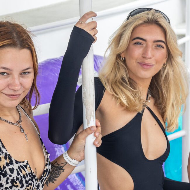 Two women are posing for a photo outdoors on a luxury yacht in Sydney. The woman on the left has auburn hair, wears a leopard print top, and has a tattoo on her upper arm. The woman on the right has blonde hair and wears a black cut-out swimsuit. Both are smiling and holding a white pole.