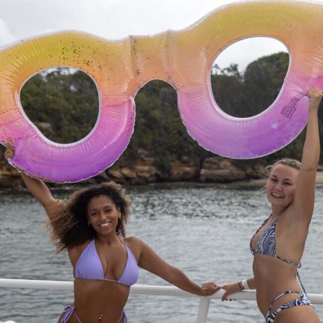 Two women in swimsuits stand on a boat, smiling and holding up an inflatable pool float shaped like oversized sunglasses with a gradient of yellow to purple. In the background, a picturesque rocky shoreline with lush trees and calm water creates a serene atmosphere.