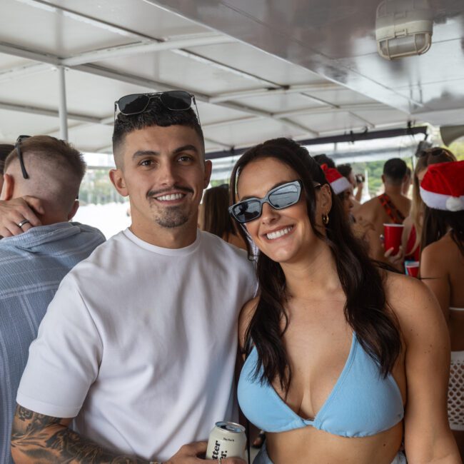 A man and woman smiling on a boat. The man is wearing a white shirt with sunglasses on his head, and the woman is in a light blue bikini top with dark sunglasses. They are surrounded by others in festive attire, enjoying beverages together as they bask in the joy of celebration.