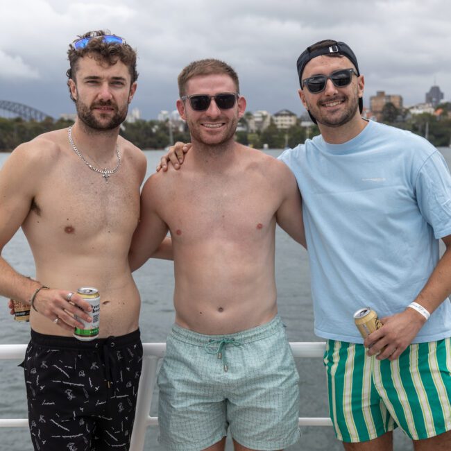 Three men stand on a boat with water and a distant shoreline in the background. They are smiling, wearing sunglasses, and casual summer attire. The man on the left and right hold drinks, while the man in the middle stands shirtless with his arms around the others.