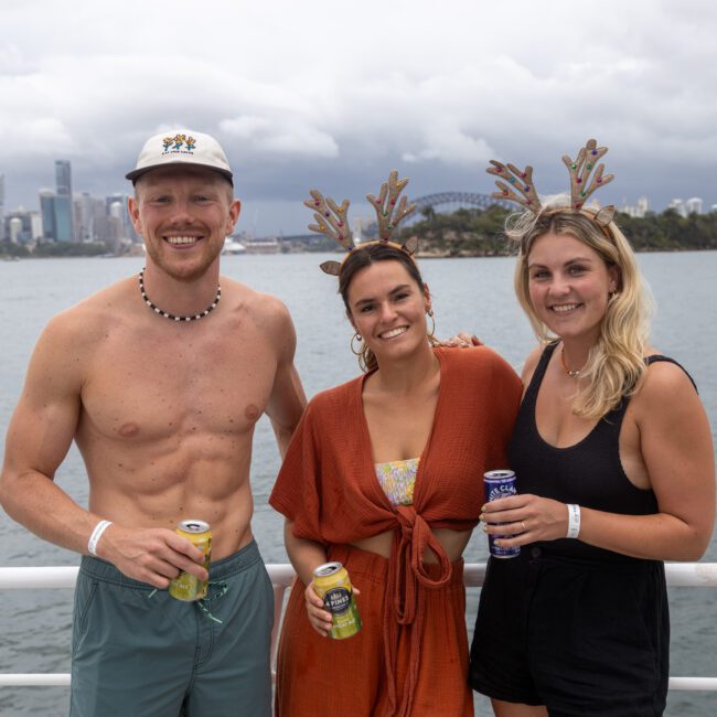 Three people stand on a boat holding drinks and smiling in front of a city skyline. The shirtless man on the left is wearing a necklace and cap, while the two women beside him are both wearing festive headbands with antlers. The sky is cloudy, with water shimmering in the background.