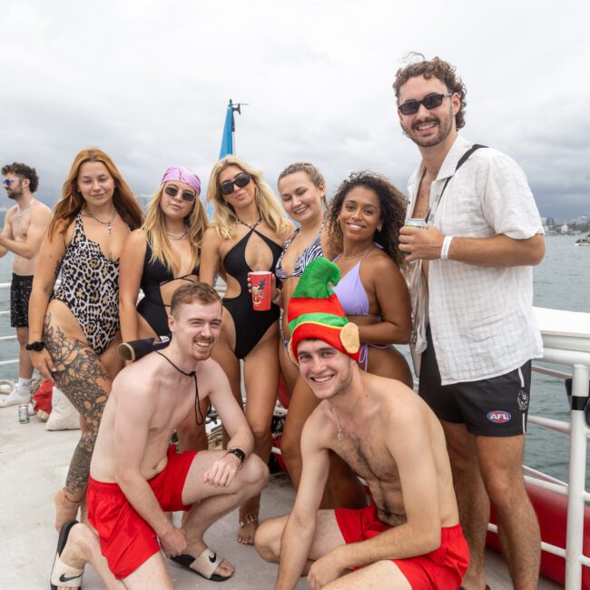 A group of people stands closely together on a boat deck, smiling at the camera. They are dressed in swimsuits, with one person sporting a green and red holiday hat. The backdrop reveals a vast body of water with a city skyline under a cloudy sky, creating an inviting atmosphere for adventure.