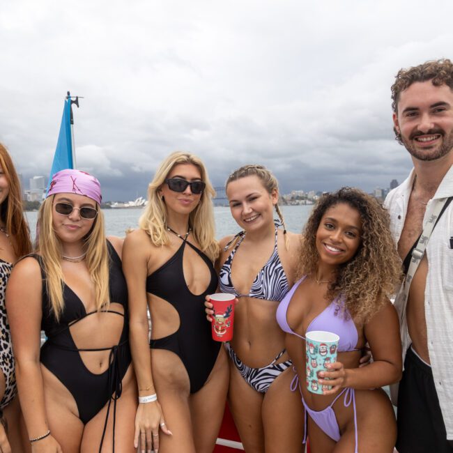 A group of five women and one man stand close together on a boat with a body of water and a cloudy sky in the background. The women wear various bikinis and swimsuits, while the man wears a light-colored shirt. Some hold cups adorned with tropical patterns, and they all smile at the camera, enjoying their day.