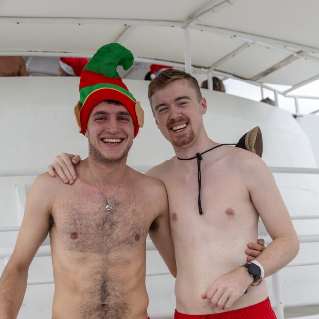 Two smiling shirtless men stand side by side on a boat. One is wearing a festive green and red elf hat while the other has a strap around his neck. The background shows the boat's structure and railing, creating a relaxed and cheerful atmosphere with an added touch of holiday fun.