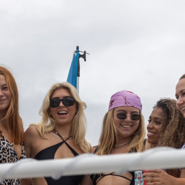 Five women smiling and posing together on a boat. They are wearing swimsuits, with one woman in a purple bandana, and another with sunglasses. In the background, there is a flagpole with a blue piece of fabric fluttering gently. The cloudy sky adds a serene ambiance to their joyful moment.