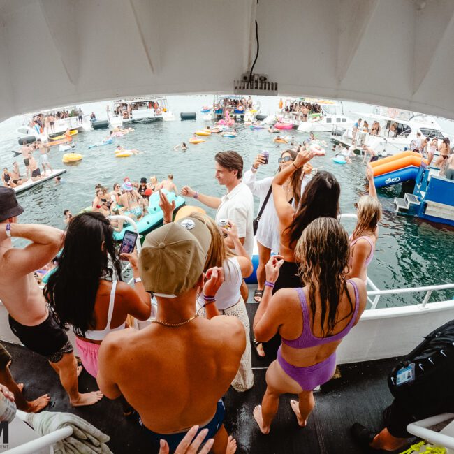 A lively crowd of people in swimwear are on a boat deck, enjoying a party with drinks and socializing. Various colorful kayaks and floaties are in the water, with more people gathered around. The scene conveys a vibrant, festive atmosphere under the clear blue sky.