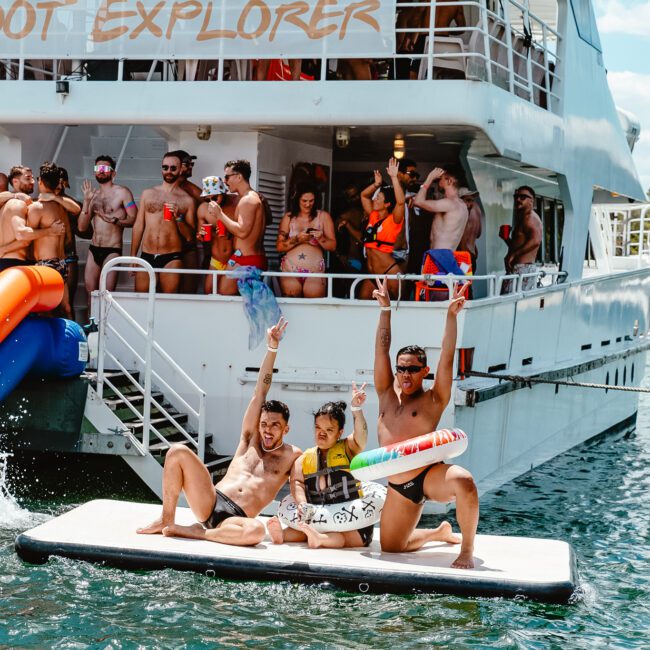 A lively scene on a boat with people in swimsuits enjoying a party. Some individuals are on an inflatable raft next to the boat, making celebratory gestures under the sun. The boat has "Barefoot Explorer" written on it, and the water below is calm and inviting.