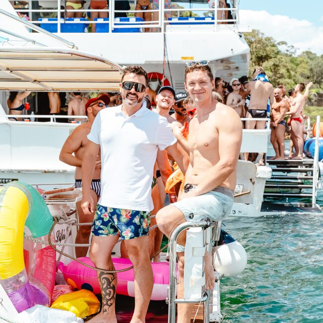 A lively boat party with people in swimwear relaxing and socializing on a sunny day. Two men are in the foreground, one in a white polo shirt and patterned shorts, and the other shirtless with turquoise shorts, standing near colorful beach balls on the water.