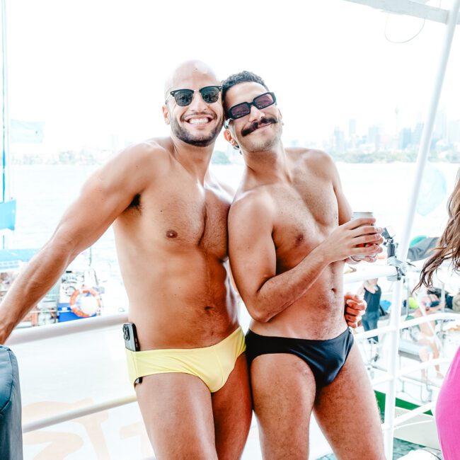 Two men smiling and posing shirtless on a boat with a city skyline in the background. One man is wearing yellow swim briefs and sunglasses, while the other wears black swim briefs and holds a can. The boat deck displays various festive banners, enhancing the vibrant atmosphere.