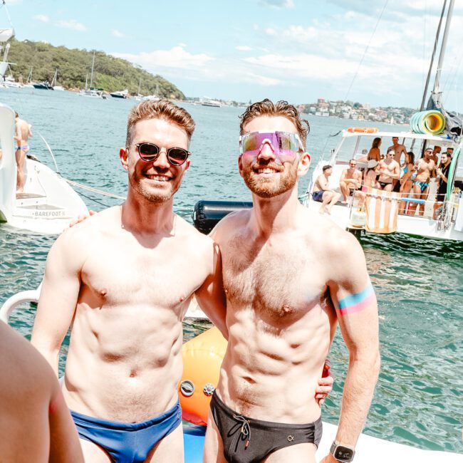 Two men wearing swim briefs stand on a boat, smiling under a sunny sky. Surrounded by water and other boats, one has his arm over the other's shoulder in a celebratory atmosphere. People on nearby boats enhance the sense of festivity and fun.