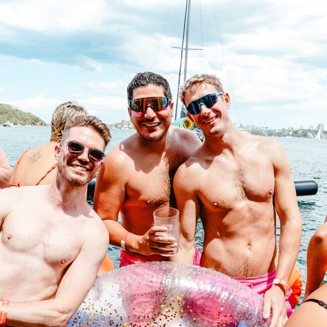 A group of shirtless individuals smiling and posing on a boat on a sunny day. Three men in the center wear sunglasses and have their arms around each other, one holding a drink. An inflatable float is visible in the foreground, with a stunning cityscape and waterway forming the backdrop.