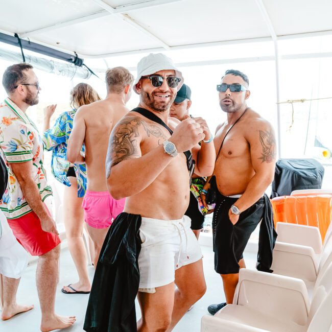 A group of people is socializing on a boat wearing casual summer attire. Two men in the foreground, sporting sunglasses, white bucket hats, and swim trunks, strike a pose. Other attendees in fashionable summer outfits are mingling and enjoying the scene in the background.