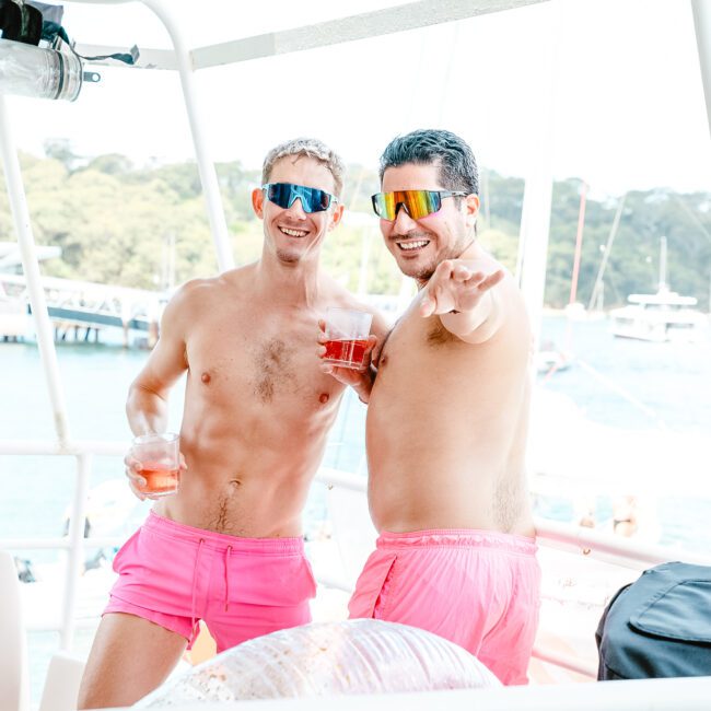 Two men in matching hot pink shorts and mirrored sunglasses are smiling and posing on a boat. One is holding a drink while the other points towards the camera. The vibrant background showcases water, boats, and lush greenery, capturing a lively summer vibe.