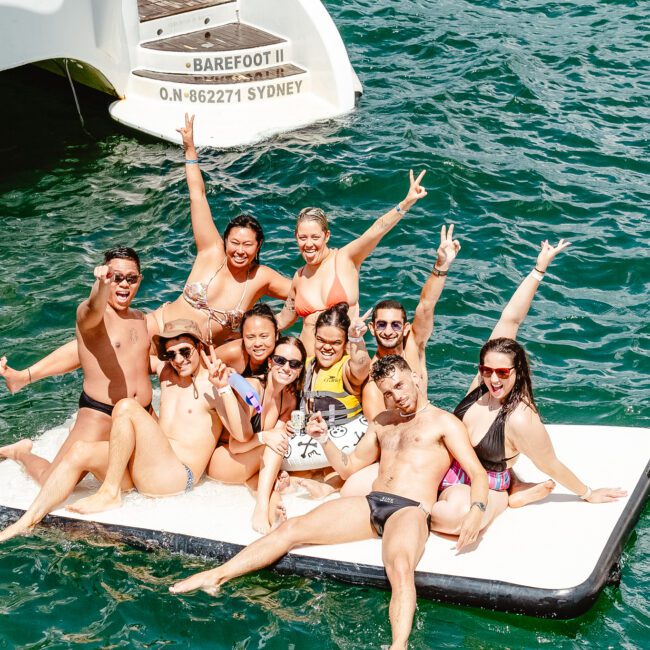 A group of people are enjoying their time on a floating platform in the water next to a boat. Clad in swimsuits and grinning widely, they pose with peace signs. The water is a vibrant blue-green, and the boat named "Barefoot III" is partially visible in the background, adding to their delightful aquatic adventure.