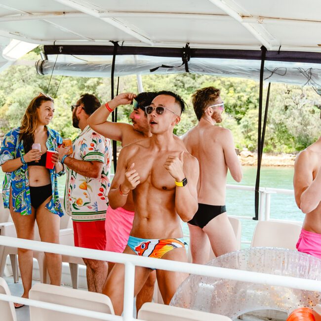 A group of people are enjoying a sunny day on a boat, wearing colorful swimsuits and sunglasses. Some are drinking and chatting, while others pose for photos. The boat sails on crystal-clear blue water with lush green foliage in the background. The scene is lively and fun-filled.