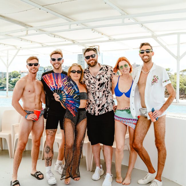 A group of six people are standing together on a boat deck. They are dressed in summer attire including swimsuits, sunglasses, and casual clothing. Some of them are holding accessories like fans. The background shows buildings and trees under a clear blue sky.