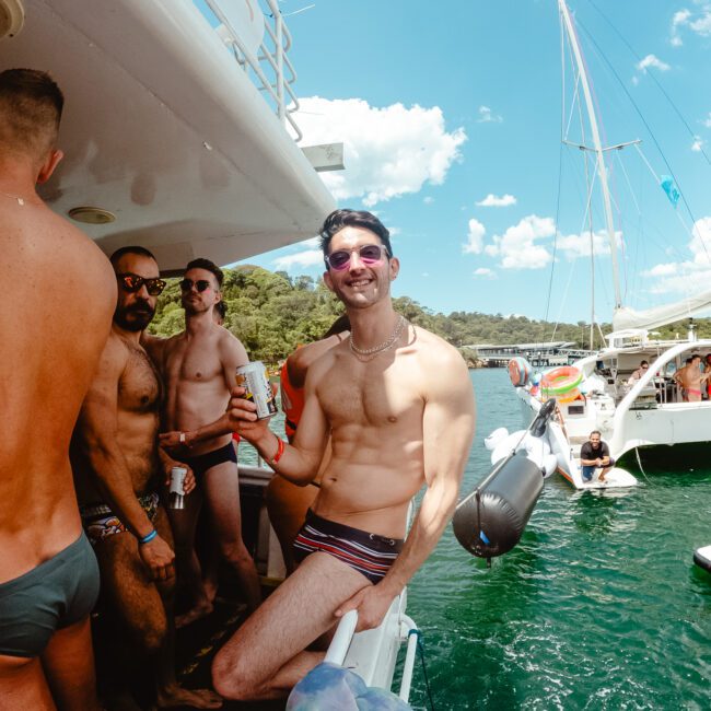 A group of men in swimwear enjoying a lively boat party on a sunny day. One man in the foreground is leaning on the boat's railing, smiling and holding a can. Other boats and people are visible in the water around them, with a clear blue sky and lush greenery enhancing the vibrant atmosphere.