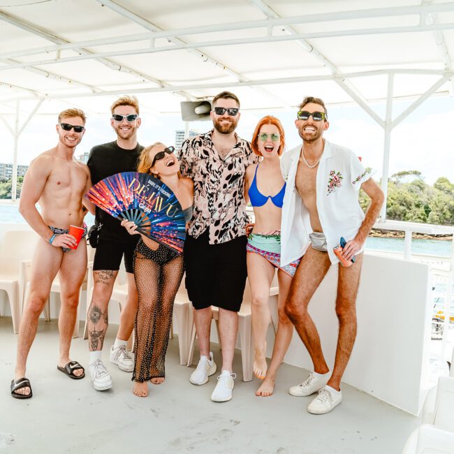 A group of six people posing on a boat deck with a scenic water view in the background. Some are wearing swimwear, with one holding a vibrant fan. They appear to be enjoying a sunny day and have a fun, relaxed vibe that perfectly captures the essence of summer adventure.