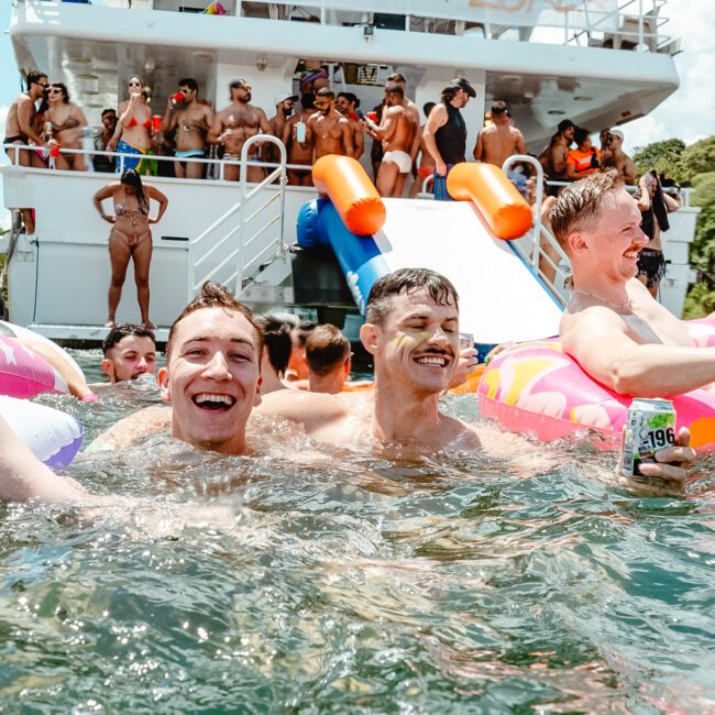 A group of people enjoying a sunny day on the water with colorful inflatable floats and a boat in the background. Some are in the water while others are on the boat, which has an exciting slide leading into the water. Everyone appears to be having fun and smiling.