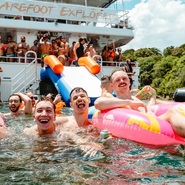 A group of people enjoys a sunny day in the water with inflatable floats near a large boat named "Barefoot Explorer." Some are on the boat's deck, while others are in the water smiling and holding drinks. Trees and a pristine blue sky create a picturesque background.