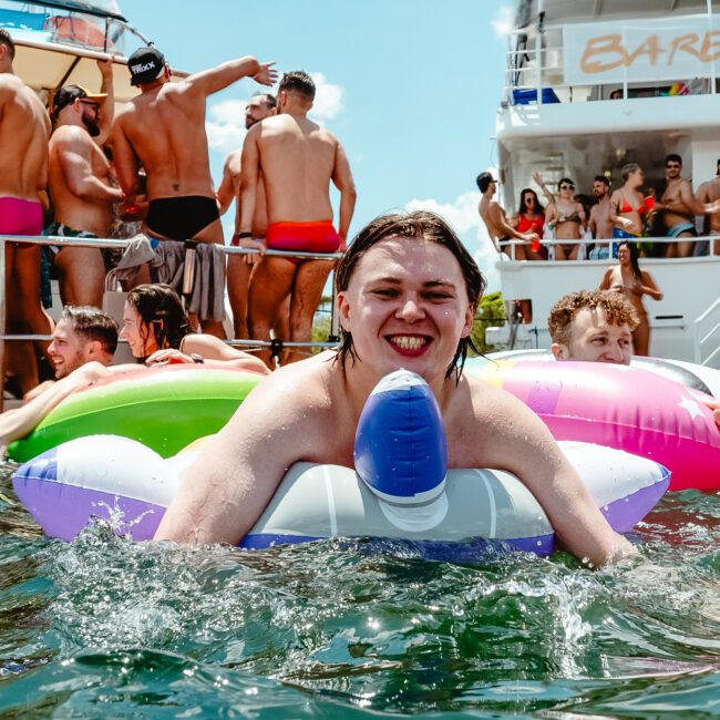 A person smiles while floating on a colorful inflatable in the water, surrounded by others in swimwear. Behind them, a party boat named "BAREFOOT" is docked, with more individuals on board enjoying the lively festivities. The sky is clear and sunny, adding to the joyous atmosphere.