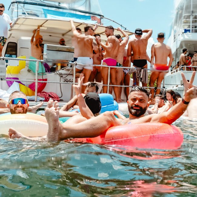 A festive boat party with many people in swimsuits on a boat. Two men in colorful swim trunks and sunglasses float on inflatable flamingos, smiling and holding up peace signs. The sunny atmosphere is lively and fun.