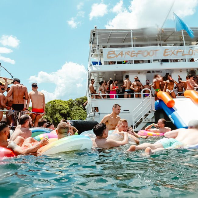 A group of people enjoying a lively day on the water with floats and inflatables. Some are in the water while others are on a boat labeled "Barefoot Explorer." The scene is bright and festive, with lush greenery and playful dolphins in the background.