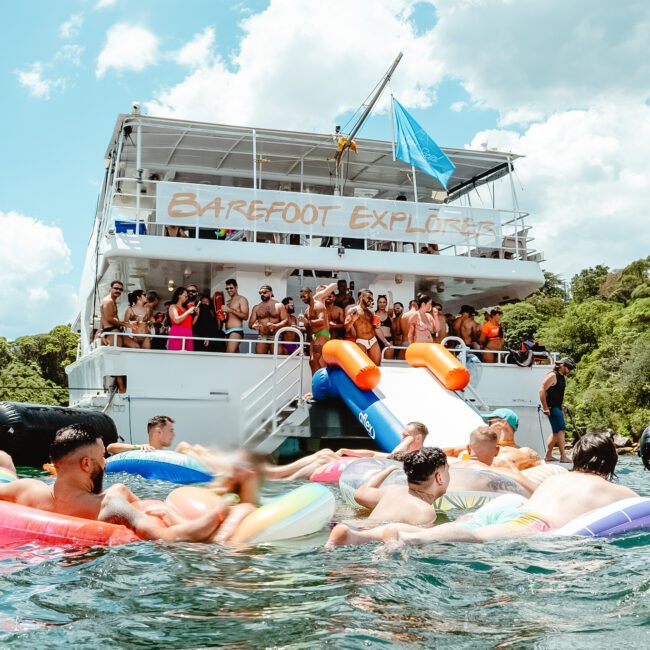 A lively scene of people enjoying a sunny day on and around a large boat named "Barefoot Explorers." Some are on the boat, while others are in the water, using colorful inflatables and sliding down giant slides. The sky is clear with a few clouds, creating an idyllic backdrop for this aquatic adventure.