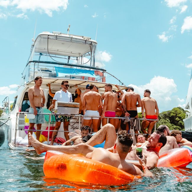 A group of people are enjoying a sunny day on a yacht. Some are lounging on colorful inflatables in the water while others are gathered on the boat, chatting and laughing. The sky is blue with a few clouds. A logo with "GAYM" is visible in the bottom left corner of the image, enhancing its appeal.