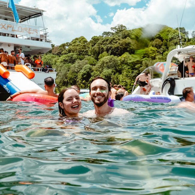 A group of people enjoy a sunny day on the water with boats and inflatables. In the foreground, a smiling couple is in the water, embracing joyfully. Behind them, others are seen socializing and having fun on the boats while vibrant birds soar above. Lush greenery is visible in the background.