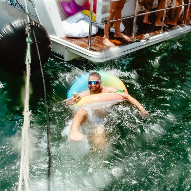 A person wearing sunglasses floats on an inflatable pool ring in the water near a boat, smiling. The boat has several people on board, with legs visible, and is docked. The image captures a sunny day with vibrant activity around the boat and sparkling water.