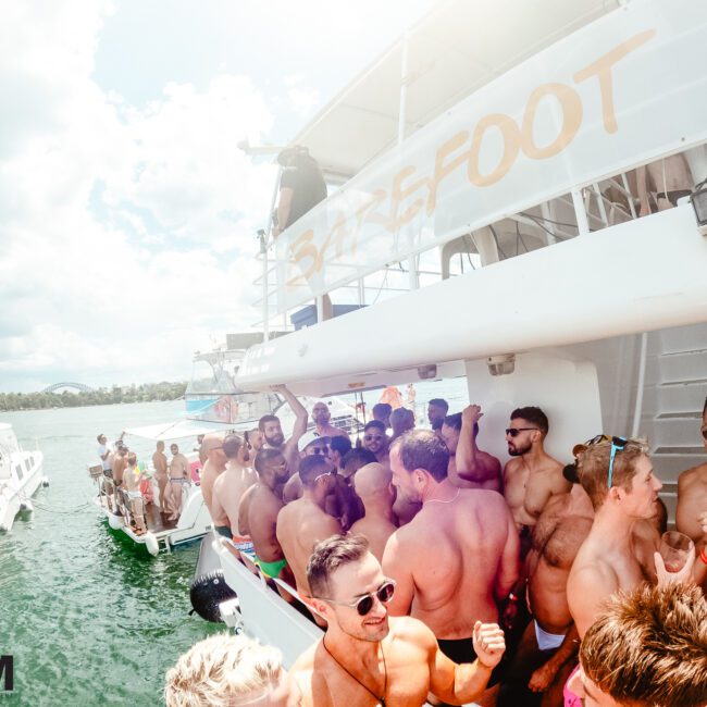 A large group of people, mostly men, are congregated on a boat labelled "BAREFOOT," with many shirtless and wearing sunglasses. Two other boats are visible in the sparkling water nearby. The partly cloudy sky adds to the festive and lively atmosphere of this outdoor gathering.