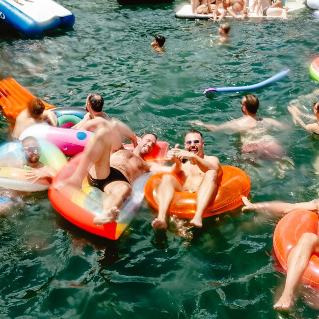 A lively group of people enjoy a sunny day on the water, floating on colorful inflatables and inner tubes. Some relax, while others engage in playful antics. The radiant scene is joyful, encapsulating the essence of a fun outdoor gathering with friends.