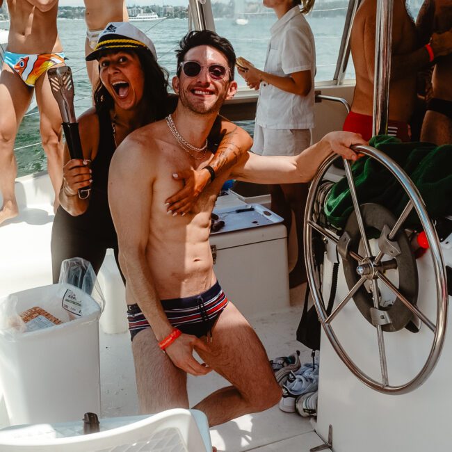 A group of people enjoy a sunny day on a boat. A man in striped swim trunks, sunglasses, and a captain’s hat smiles while playfully having his chest touched by a woman in another captain’s hat. Other people in swimwear are relaxing, mingling, and savoring the refreshing sea breeze in the background.