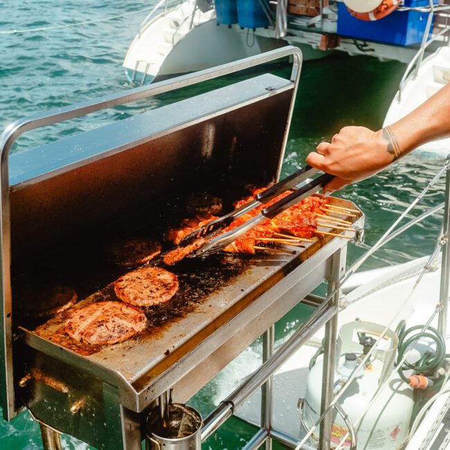 A barbecue grill on a boat with burgers and skewers being cooked. A person's hand with a spatula is seen flipping the food, adding to the nautical excitement. The background shows a body of water and another boat. The GAYM logo is visible in the lower-left corner.