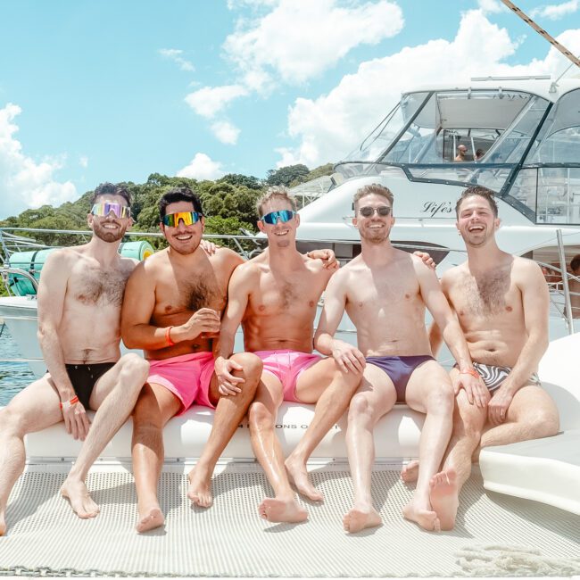 Five men in colorful swim trunks are sitting and smiling on a catamaran's netting. Behind them, a boat with another group of people is docked. The backdrop features lush green trees and a partly cloudy sky, creating a picturesque scene perfect for any sailing enthusiast.