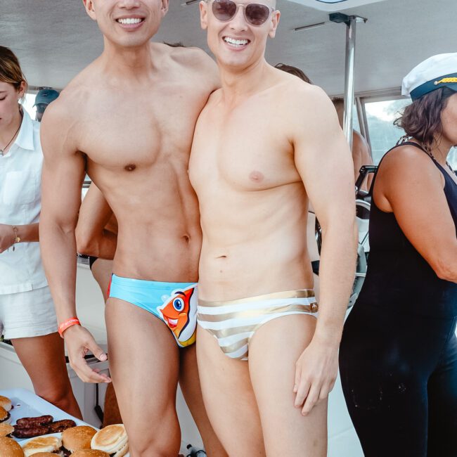 Two men stand smiling on a boat, wearing colorful swim trunks. One sports a blue swimsuit with a fish design, the other dons a striped white and beige swimsuit. There are other people in the background and a table brimming with delicious food. The boat's windows offer a scenic backdrop.