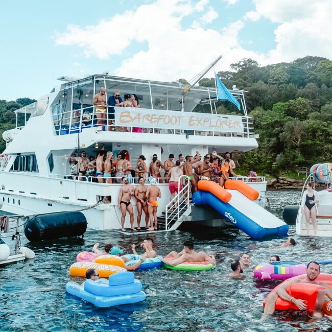 A lively scene of people partying on a large white boat named "Barefoot Explorer" and in the water around it. Some are on the boat's deck, while others enjoy inflatable floats and slides into the water. The backdrop features lush tropical trees under a partly cloudy sky, creating an idyllic summer vibe.