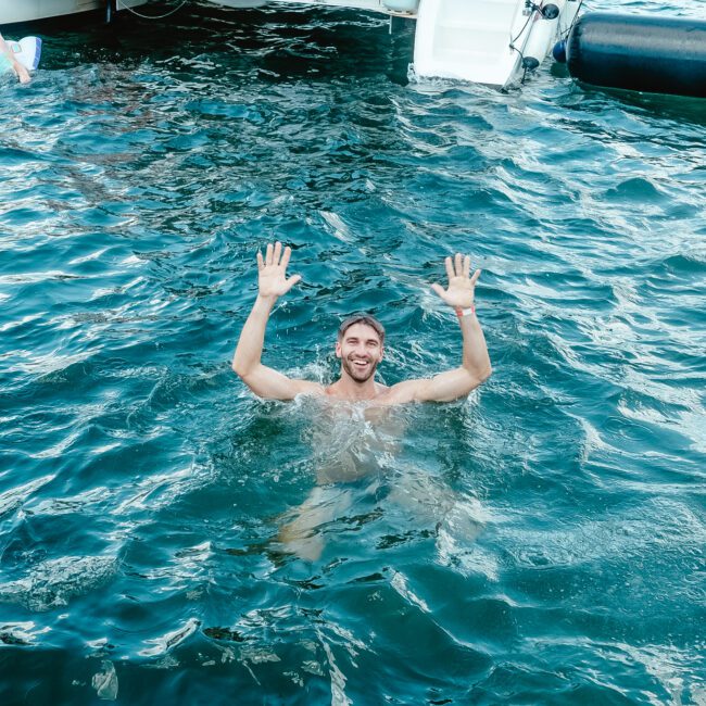 A man standing waist-deep in the clear water with his hands raised, smiling at the camera. A scenic boat is visible in the background, with a few people on board enjoying the day.