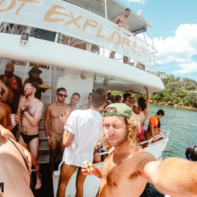 A group of people are enjoying a sunny day on a boat named "ECOT EXPLORER". The crowd appears to be socializing, with some holding drinks. In the foreground, someone is taking a selfie, squinting their eyes with a slight smile. Water and lush green trees are visible in the background.