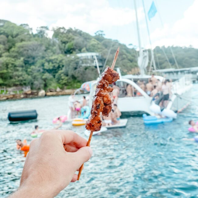 A hand holding a skewer of grilled meat is in focus, with a lively scene of people enjoying a festive gathering on boats in the background. The waters are clear, and the boats are equipped with inflatable slides and floats. A lush, green shoreline completes the picturesque backdrop.