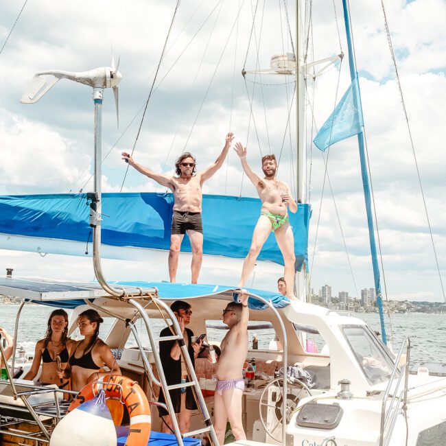 A group of people on a sailboat enjoys a sunny day. Two individuals stand on the top deck holding hands, raising their arms in celebration, while others are seated or standing on the boat's main deck. The backdrop features sparkling water and a distant city skyline.