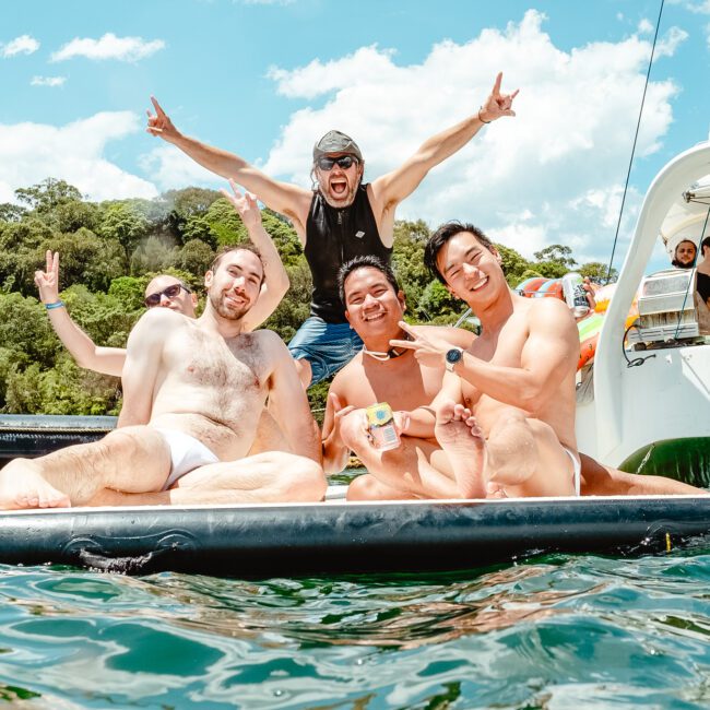 A group of four men relaxes on an inflatable float in a body of water, with one man behind them raising his arms enthusiastically. A boat with more people is in the background. The scene is bright and sunny, suggesting a fun, recreational day featuring water sports activities.