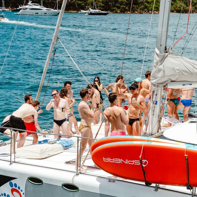 A group of people in swimwear socialize and enjoy themselves on a luxurious yacht anchored near a scenic shoreline. The yacht features a paddleboard and a shaded cockpit area. The bay is dotted with other boats, and the water is calm and blue. Trees line the distant shore, adding to the serene ambiance.