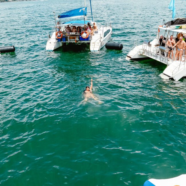 A swimmer floats in clear blue water alongside three catamarans filled with people enjoying a sunny day. Some boats have vibrant red flags, and passengers on the decks relax and engage in recreational activities like snorkeling. The festive scene unfolds against a backdrop of the open sea.