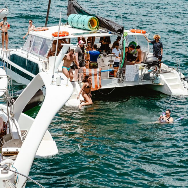 A group of people enjoying a sunny day on two connected boats at sea. Some are lounging on the boats, while others are swimming or snorkeling in the clear, blue water. Towels and other beach items are visible on the boats, creating a perfect seaside getaway.