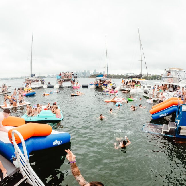 A lively boat party on a lake with numerous people swimming and lounging on colorful inflatables. Several boats are anchored close together, and the water is filled with partygoers enjoying the festivities of this lakeside celebration. The sky is overcast, creating a relaxed ambiance perfect for fun.